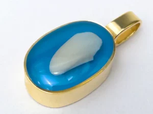 Ein goldener Schmuckanhänger mit einem auf blauem Grund in klares Kunstharz eingegossenen Hundezahn.